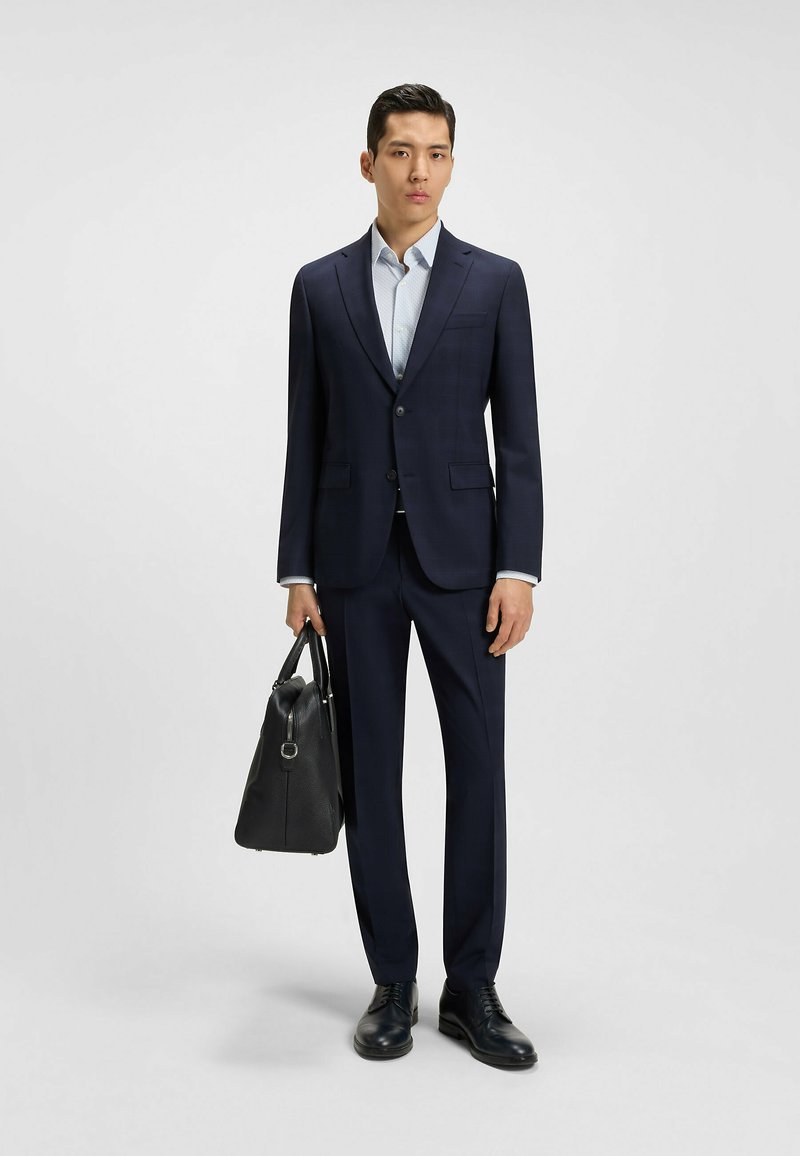 Marinblå kostym med smal passform, en knapp och subtil rutmönstring. Ljusblå skjorta, svart läderportfölj och polerade skor.