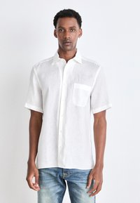 Camisa de lino blanca de manga corta con cierre de botones y un único bolsillo en el pecho, combinada con jeans de mezclilla azul claros sobre un fondo liso.