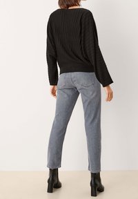 Osoba ubrana w prążkowany czarny sweter, jasnoszare dżinsy z wysokim stanem i czarne botki na obcasie, stojąca w pobliżu gładkiej ściany.