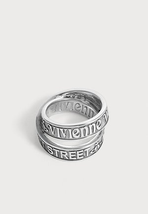Sølv ring, der omslutter fingeren, med præget tekst "Vivienne Westwood" og "STREET 37" på en ensfarvet lys baggrund.