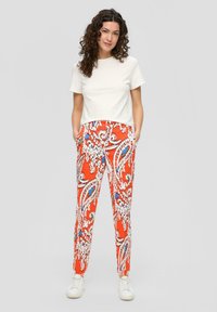 Pantalon à motif cachemire rouge avec des accents bleus et blancs, associé à une chemise blanche à manches courtes unie. Le mannequin se tient avec les mains dans les poches.