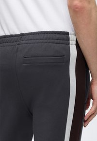 Gros plan d'une personne portant un pantalon gris foncé avec des bandes latérales blanches et marron et une poche passepoilée arrière, associé à une chemise blanche.