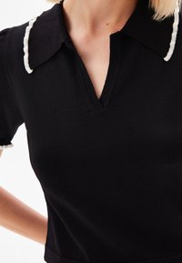 Black knit polo shirt featuring a deep V-neckline, short puff sleeves, and a white, ribbed collar detail. Smooth texture and fitted design.