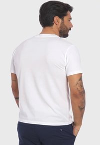 T-shirt bianca a maniche corte, design con colletto girocollo. Tessuto liscio, cuciture minime. Indossata da una persona con capelli corti scuri e tatuaggi sul braccio destro.