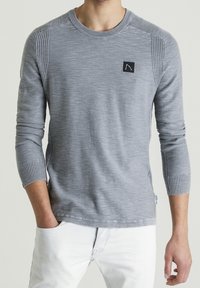 Maglione grigio a maniche lunghe con scollatura e polsini a costine. Presenta un piccolo patch logo nero sul petto. Tessuto morbido, design aderente.