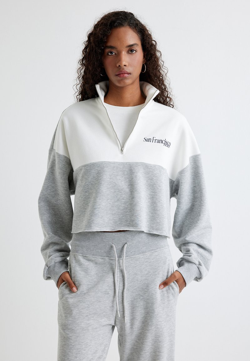 Geknipte pullover in grijs en wit, gemaakt van zacht materiaal. Heeft een hoge kraag met een halve rits en borduursel met de tekst "San Francisco."