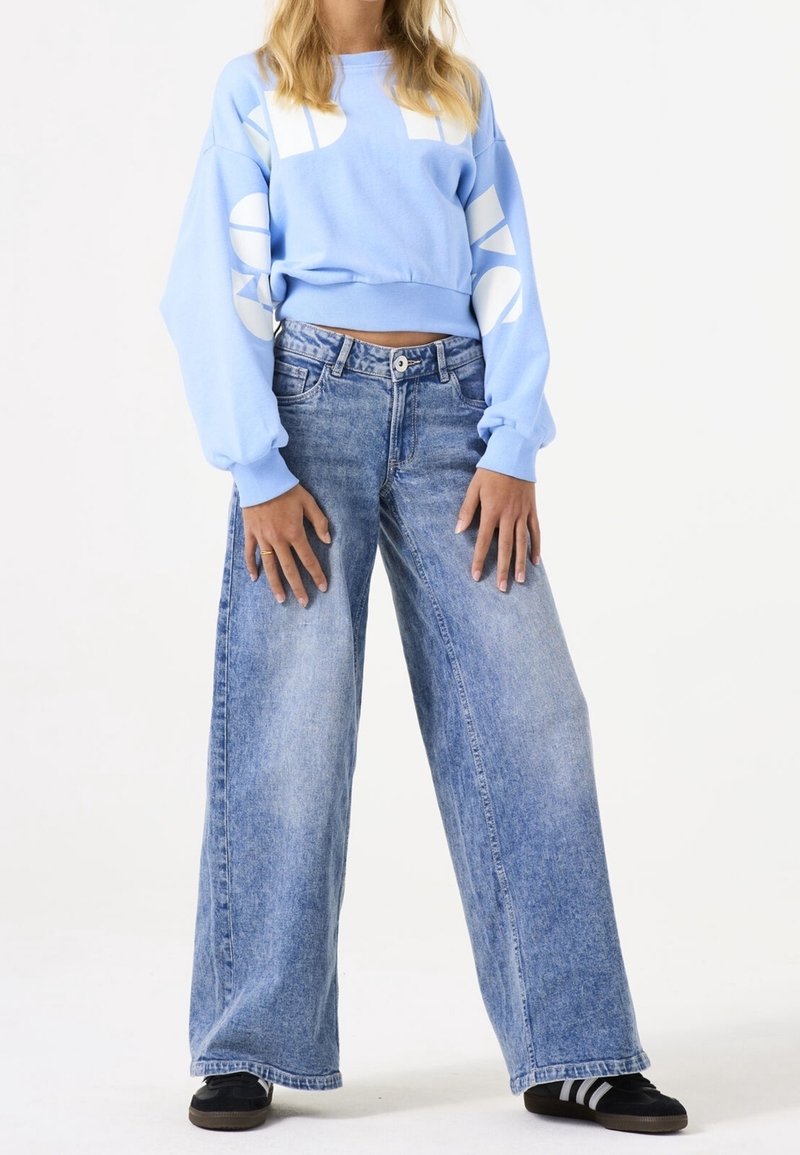Personne portant un sweat-shirt court bleu clair avec un texte blanc et un jean large bleu délavé, debout avec les mains détendues le long du corps.