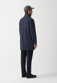 Marinejas met hoge kraag en knoopsluiting aan de achterkant, gecombineerd met donkere broeken en glanzende zwarte schoenen. Model draagt een grijze pet.