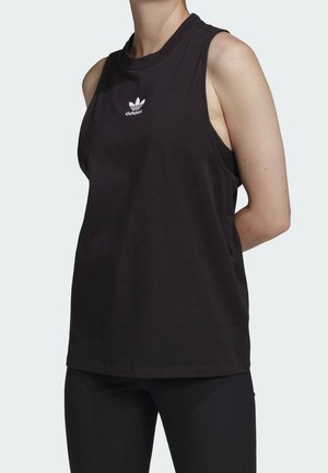 Osoba nosząca czarną bezrękawnik Adidas z logo na piersi, w połączeniu z czarnymi spodniami, stojąca na jednolitym jasnym tle.