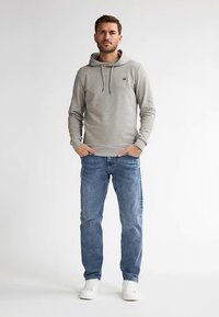 Šedá mikina s kapucí na šňůrku a logem, doplněná světle modrými džínami. Model stojí v bílých teniskách na jednobarevném pozadí.