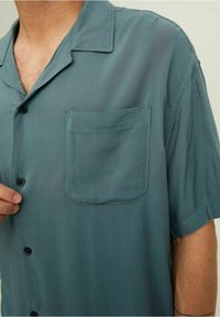 Uomo che indossa una camicia a maniche corte di colore teal con taschino, mentre tiene il bottone davanti vicino al colletto su uno sfondo neutro.