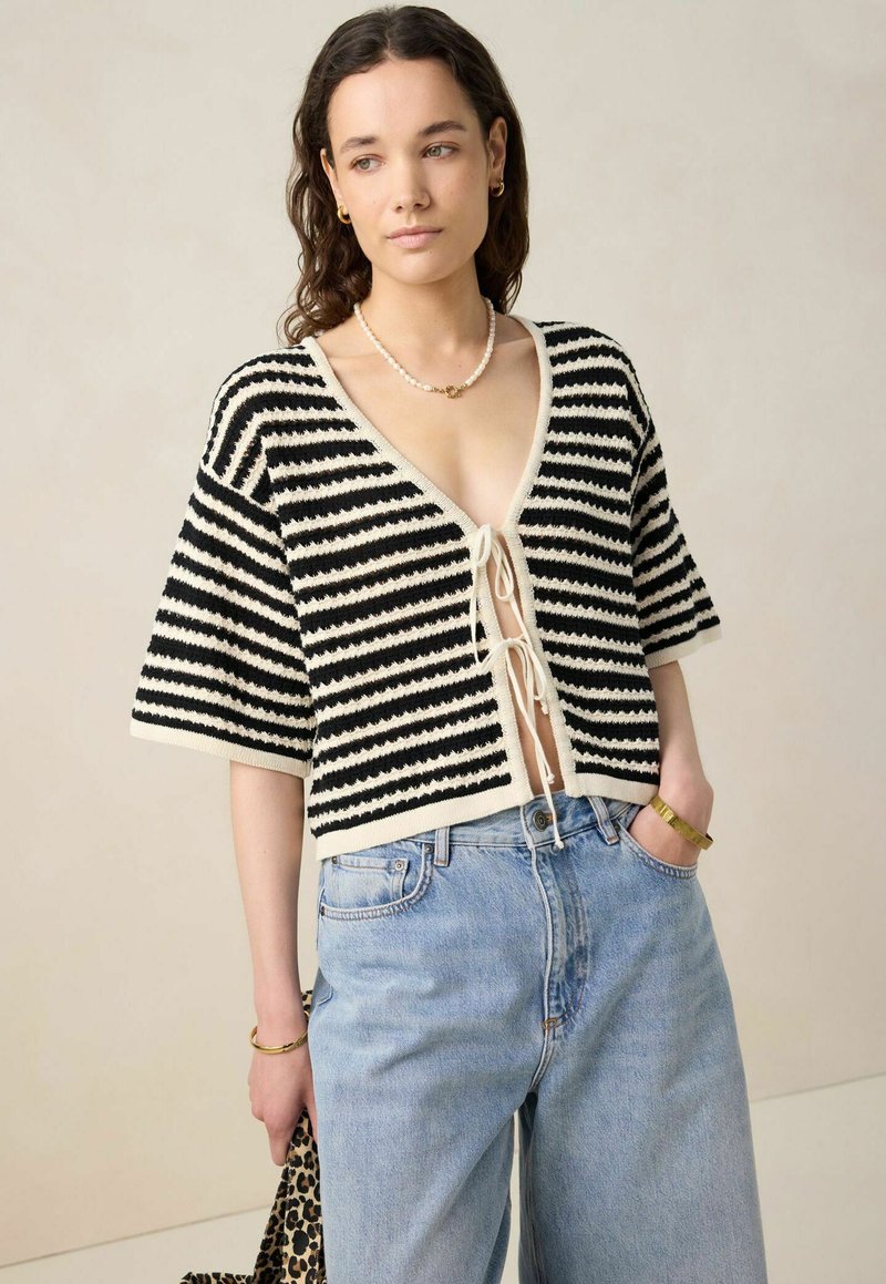 Pull cropped noir et blanc rayé avec un col en V, devant noué et manches courtes, porté avec un jean en denim bleu clair.