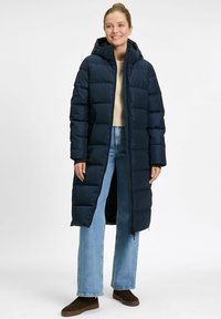 Vrouw glimlachend, gekleed in een lange marineblauwe gewatteerde jas met capuchon, beige trui, lichtblauwe jeans en bruine schoenen, staande voor een witte achtergrond.