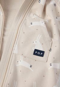 Beige zip-jacket med et mønster af hvide kaniner og brune prikker. Indeholder en lomme med en blå etiket, der står "P.O.P.". Blødt stof.