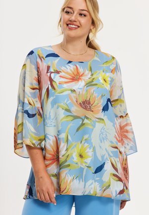 Vrouw draagt een losse bloes met bloemenpatroon in blauw, oranje, geel en groen, gecombineerd met lichtblauwe broek, ze lacht en kijkt naar de zijkant.