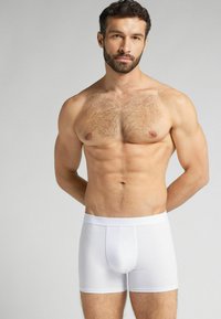 Bílé bavlněné boxerky s těsným střihem, s pasem a přední kapsou. Materiál se zdá být hladký a elastický.