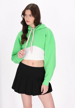 Junge Frau trägt einen grünen cropped Hoodie mit pinken Kordeln und einen schwarzen plissierten Rock, schaut zur Seite mit der Hand nahe den Hoodie-Schnüren.