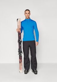 Blå jakke med glidelås, svarte bukser, holder på ski med hvite bindinger. Skiene har en gradient fra lyse til mørke brune med trykte detaljer.