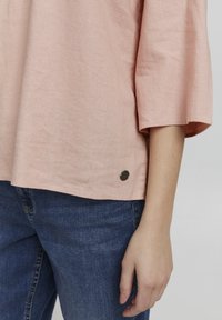 Haut rose clair, ample, en tissu texturé, avec des manches 3/4 et un petit accent métallique près de l'ourlet. Porté par-dessus un jean bleu foncé.