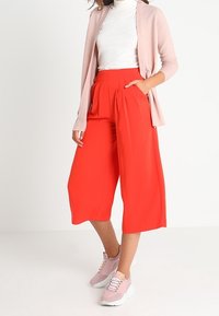 Röda culotter med veck, kombinerade med en vit ribbad polotröja och en ljusrosa kofta. Skorna består av rosa sneakers med snören.