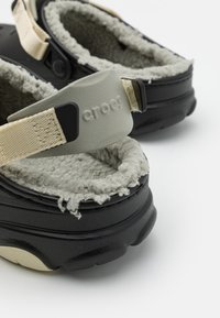 Zapatos Crocs negros con un suave forro gris de felpa, correas ajustables beige y una suela texturizada. Con un logo en la correa.