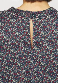 Blouse fleurie avec un fond bleu marine orné de petites fleurs rouges et blanches. Ouverture en forme de trou de serrure dans le dos, avec un col et un tissu texturé.