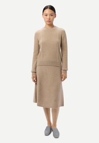 Kvinna iklädd en beige stickad tröja och matchande midikjol, stående rakt upp, med gråa slip-on skor mot en enkel vit bakgrund.