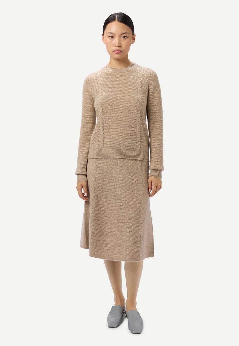 Kvinna iklädd en beige stickad tröja och matchande midikjol, stående rakt upp, med gråa slip-on skor mot en enkel vit bakgrund.