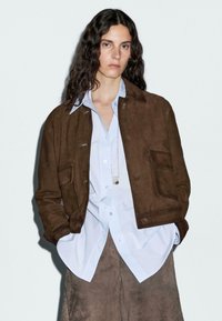 Brown suede jacket with two front pockets, collared design, worn over a light blue button-up shirt. Smooth texture, casual fit.