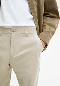Beige chinos with a smooth texture, featuring slanted front pockets and a button closure. Paired with a white t-shirt and a brown jacket.
