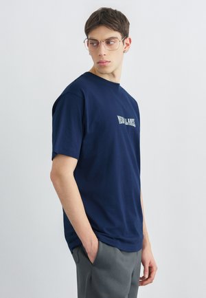 Mladý muž s krátkymi vlasmi a okuliarmi, oblečený v tmavomodrom tričku s nápisom "NEW BALANCE", ruky v vreckách šedých nohavíc, pozerá sa bokom.