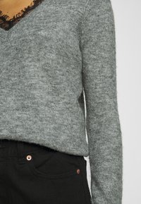 Grijze gebreide trui met een V-hals en zwarte kantafwerking, gecombineerd met zwarte jeans met koperkleurige hardware. Zachte textuur zichtbaar.