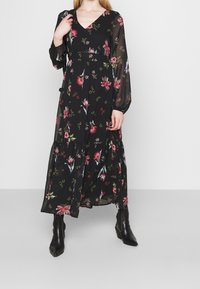 Robe maxi noire à fleurs avec des manches longues et transparentes, décolleté en V et ourlet à volants ; présente des motifs floraux rouges et verts sur l'ensemble.