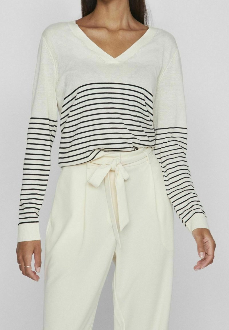 Femme portant un pull blanc à col en V avec des rayures horizontales noires et un pantalon crème taille haute noué à la taille avec une ceinture en tissu.