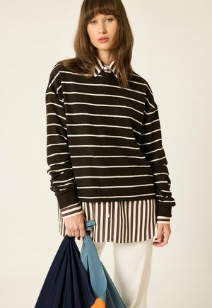 Jeune femme portant un pull rayé marron foncé et blanc par-dessus une chemise rayée marron et blanc, tenant des sacs en tissu bleus et orange.
