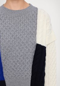 Gebreide trui met een mix van grijze, marineblauwe, blauwe en crèmekleurige panelen. Getextureerde patronen bevatten kabels en stippenweefsel; ribgebreide halslijn.