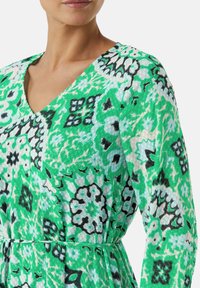 Groen gestippeld jurkje met een V-hals en lange mouwen, versierd met ingewikkelde bloemen- en geometrische patronen in zwart en aqua.