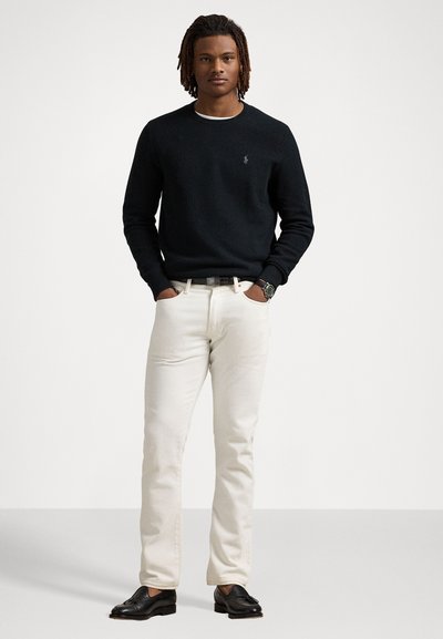 Pull en maille noir avec col rond, jeans crème et mocassins noirs. Présente un logo discret sur le pull et une ceinture sombre.