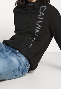 Černá mikina s kapucí vyrobená z měkké látky, která má vpředu logo v tmavě šedé barvě. Kombinována se světle modrými džíny.