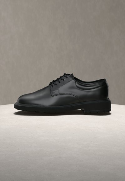 Μαύρο γυαλιστερό δερμάτινο παπούτσι derby με κορδόνια πάνω σε φωτεινή υφή επιφάνειας και μονοχρωματικό ουδέτερο φόντο.