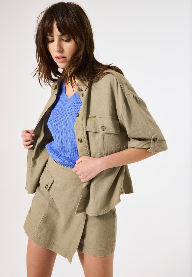 Camisa de lino color caqui con mangas enrolladas y dos bolsillos en el pecho, combinada con un suéter de canalé azul con escote en V y una falda envolvente caqui a juego.