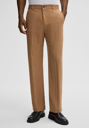Man wearing brown tailored trousers and black leather loafers, standing with one hand in pocket against plain background.