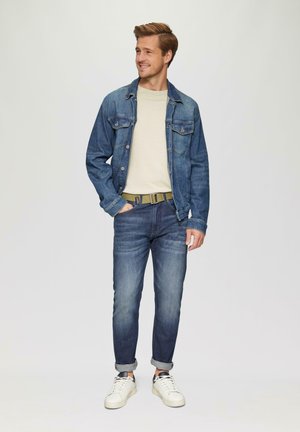 Jeansjack in medium blauw, gelaagd over een beige trui; gecombineerd met donkere spijkerbroeken en witte sneakers, met een groene riem.