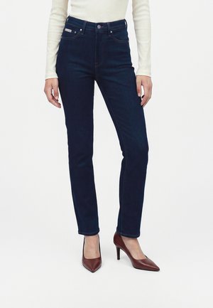 Femme portant un jean slim bleu foncé, un haut crème à manches longues et des chaussures à talons hauts bordeaux, debout devant un fond clair uni.