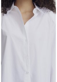 Camisa blanca de algodón ligero con botones delanteros. Cuenta con un cuello puntiagudo, botones en la parte frontal y un corte relajado con textura mínima.