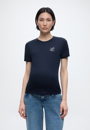 Γυναίκα με κοντά μαύρα μαλλιά που φοράει μπλε σκούρο κοντομάνικο μπλουζάκι με μικρό λευκό κείμενο και ανοιχτόχρωμο τζιν, στέκεται μπροστά σε μονόχρωμο φόντο.