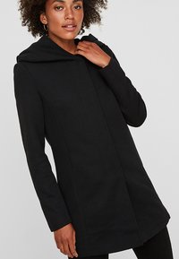 Cappotto nero con cappuccio, dalla vestibilità sartoriale, maniche lunghe e design minimalista. Realizzato in tessuto morbido, presenta una chiusura frontale nascosta.