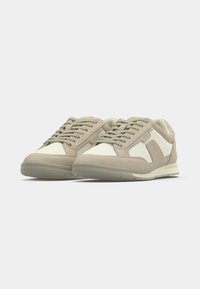 Paire de baskets basses beige et blanches avec lacets, semelles texturées et détails en panneaux de daim sur un fond uni.