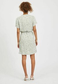 Femme aux cheveux bouclés portant une robe légère verte clair à manches courtes et à mi-longueur avec un imprimé abstrait blanc et des sandales à talons hauts blanches, se tenant de dos.