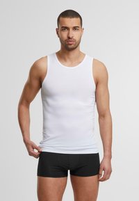 Camiseta de algodón blanca sin mangas con escote redondo, diseño ajustado, textura suave. Combinada con pantalones cortos boxers negros. El modelo está de pie con una mano en la cintura.